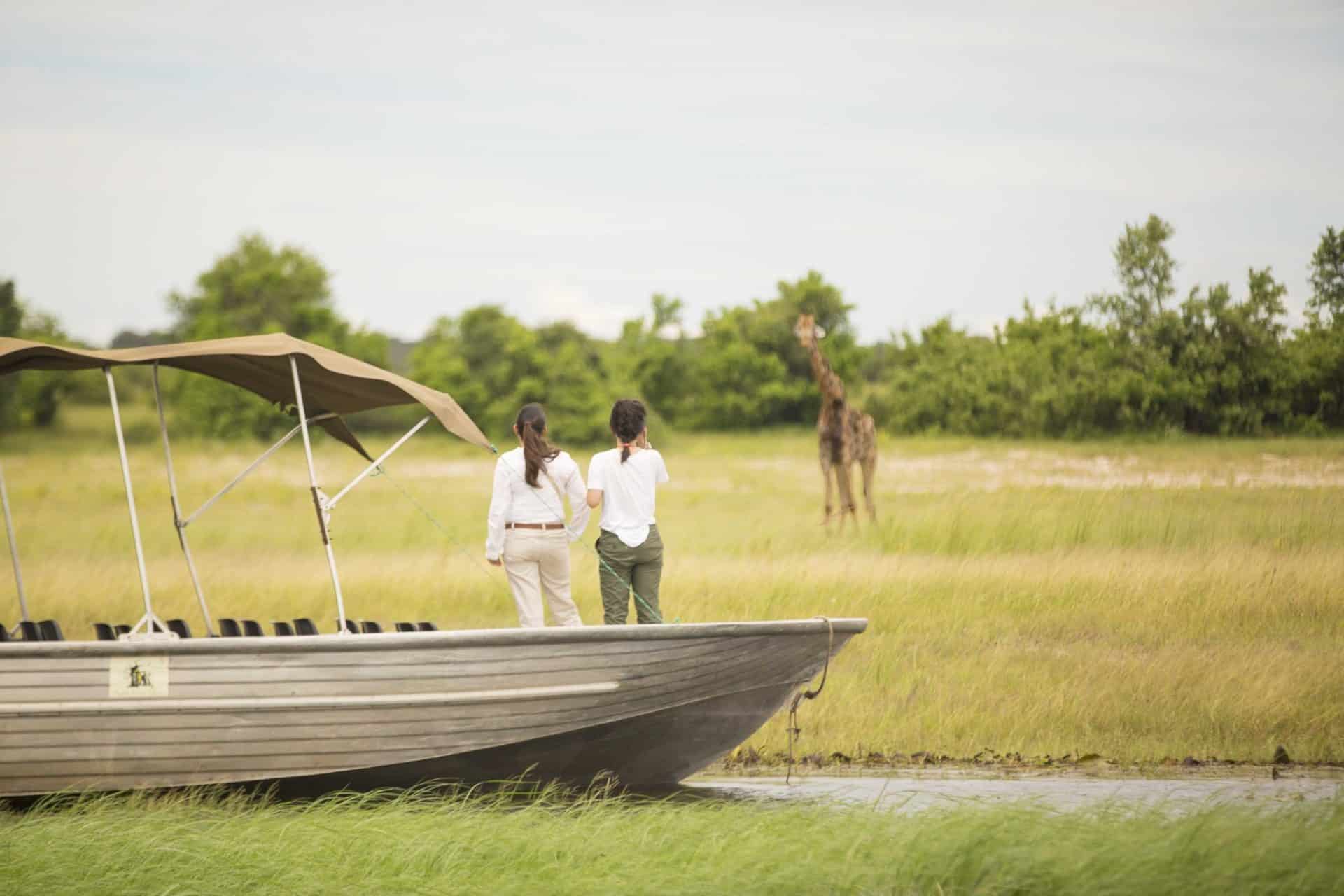 Two people observe a giraffe from a boat in Chobe National Park . Ker&Downey Africa offers the best safaris in africa to see the Big 5.