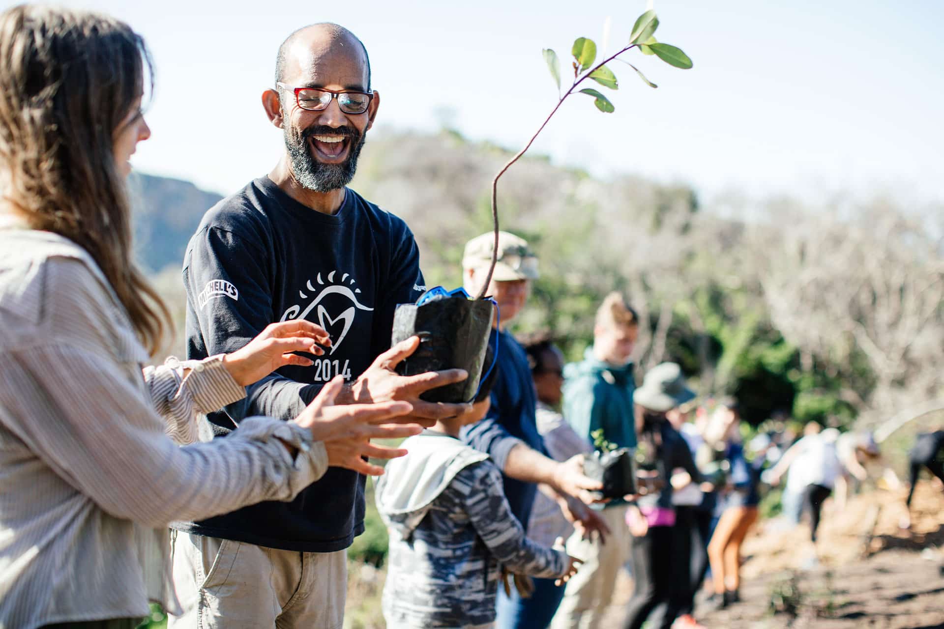 A man smiling and passing a young tree to a woman to be planted at a Greenpop planting day.