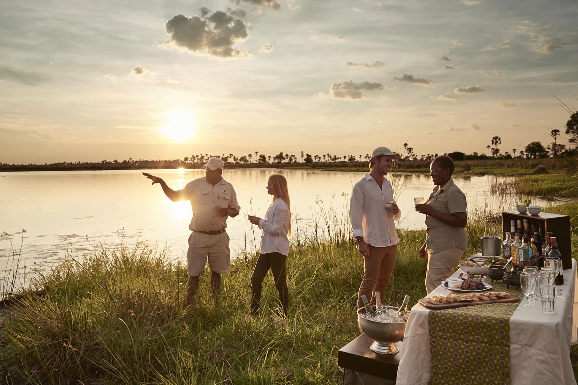 People alongside a body of water watching the sun set and having drinks before a night safari at Sanctuary Baines’ Camp.