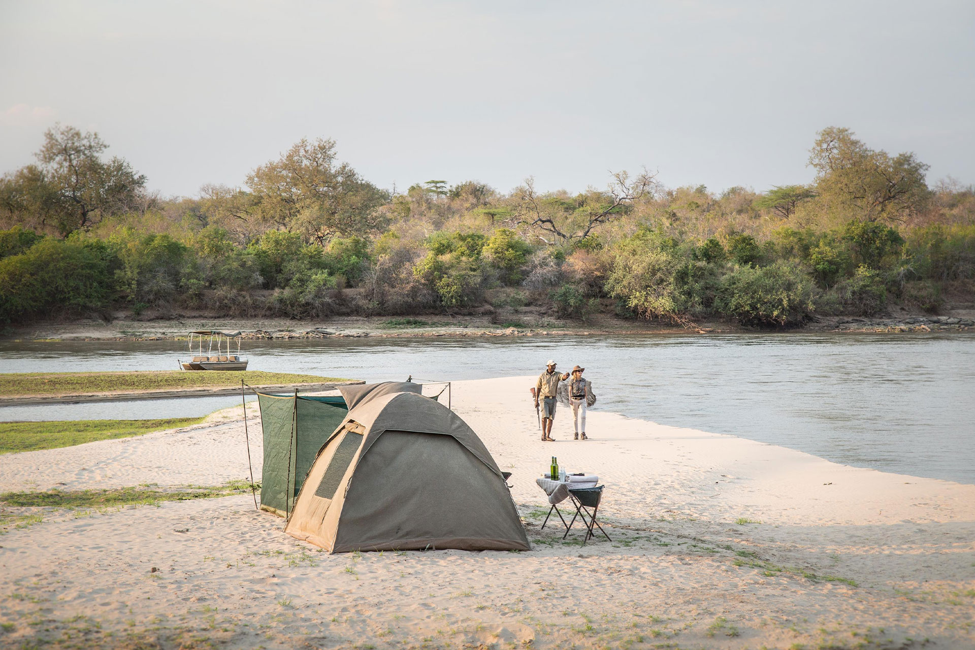 A man, woman and tent next to a river with a boat in the background on a night safari at Sands River Selous.