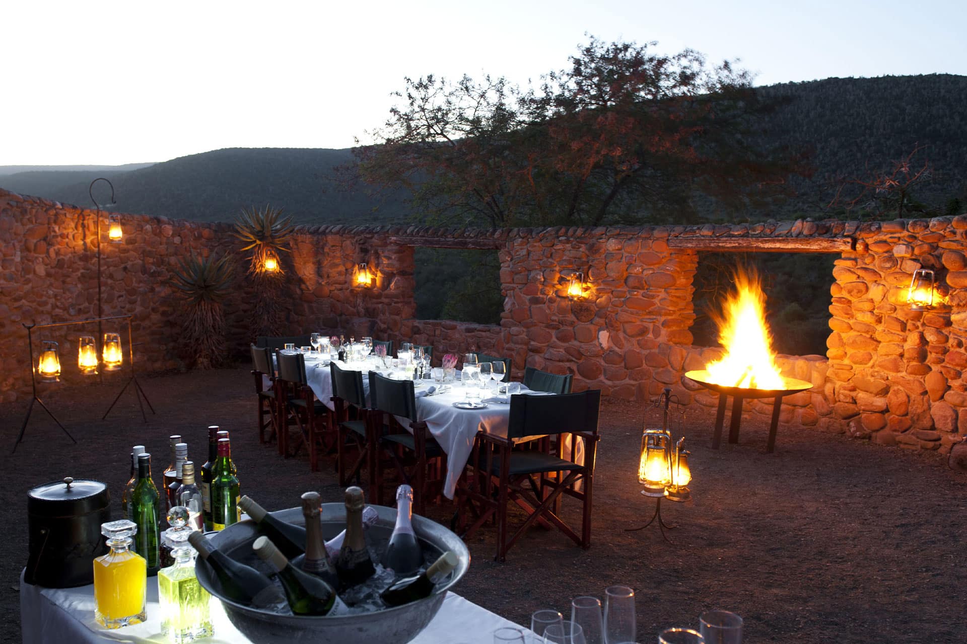 A set dinner table and drinks station in a boma with a lit campfire at Ecca Lodge at Kwandwe Private Game Reserve.