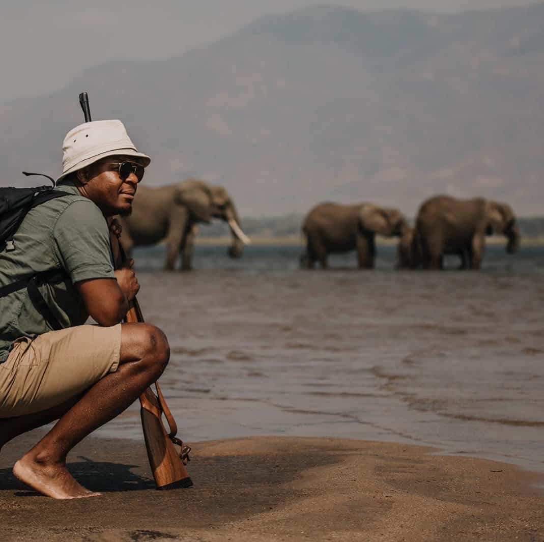 Beks Ndlovu crouching in front of a waterhole with elephants in the background.
