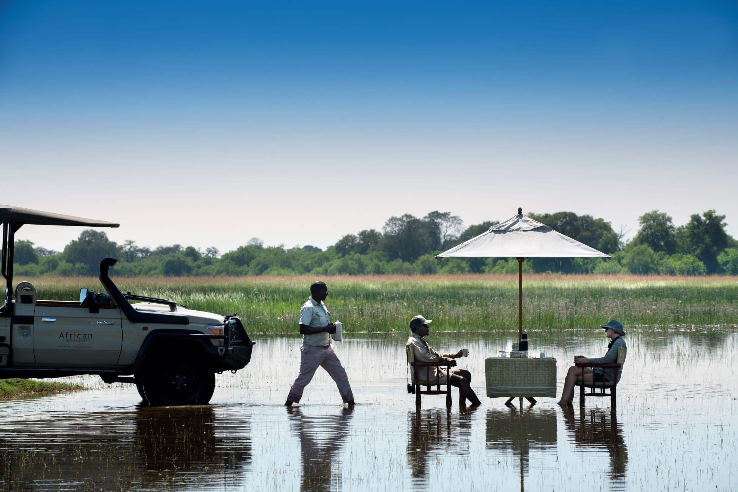 Beks Ndlovu sitting at a table being served coffee in the shallow waters of the Okavango Delta.