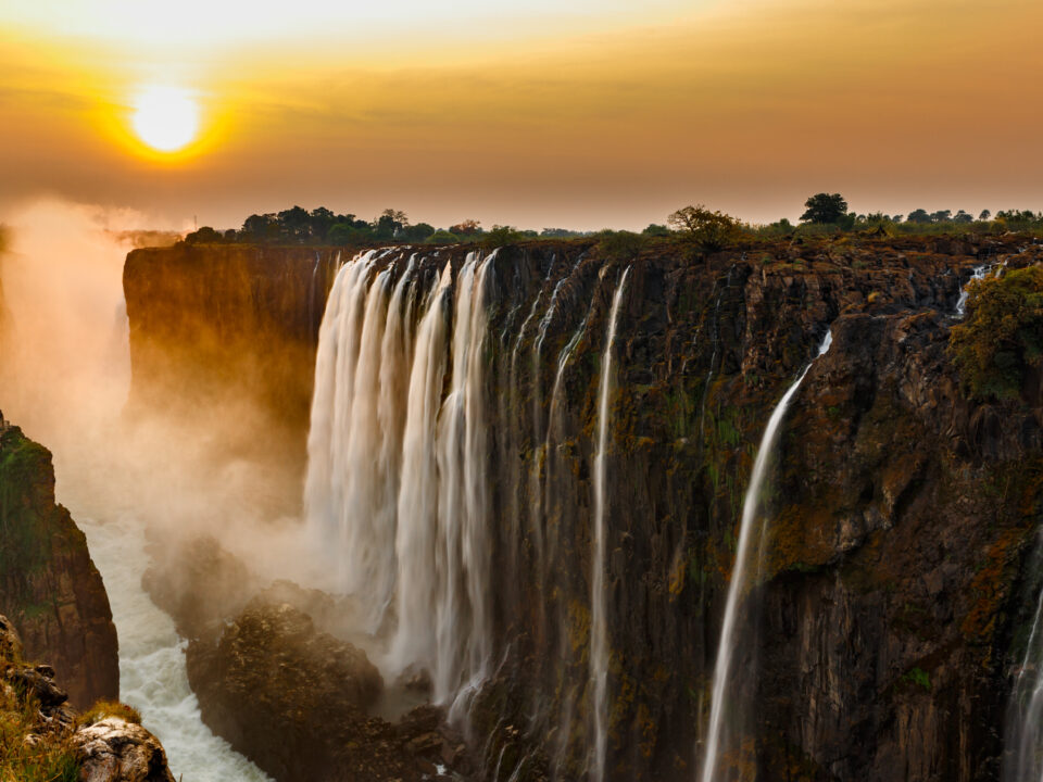 Wide panorama of victoria falls at sunset with orange sun in the sky and tourist in viewpoint