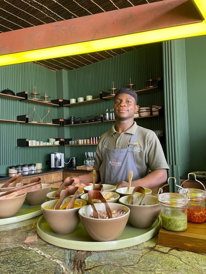 Staff member at Lolebezi with a variety of food in the front with Ker & Downey Africa