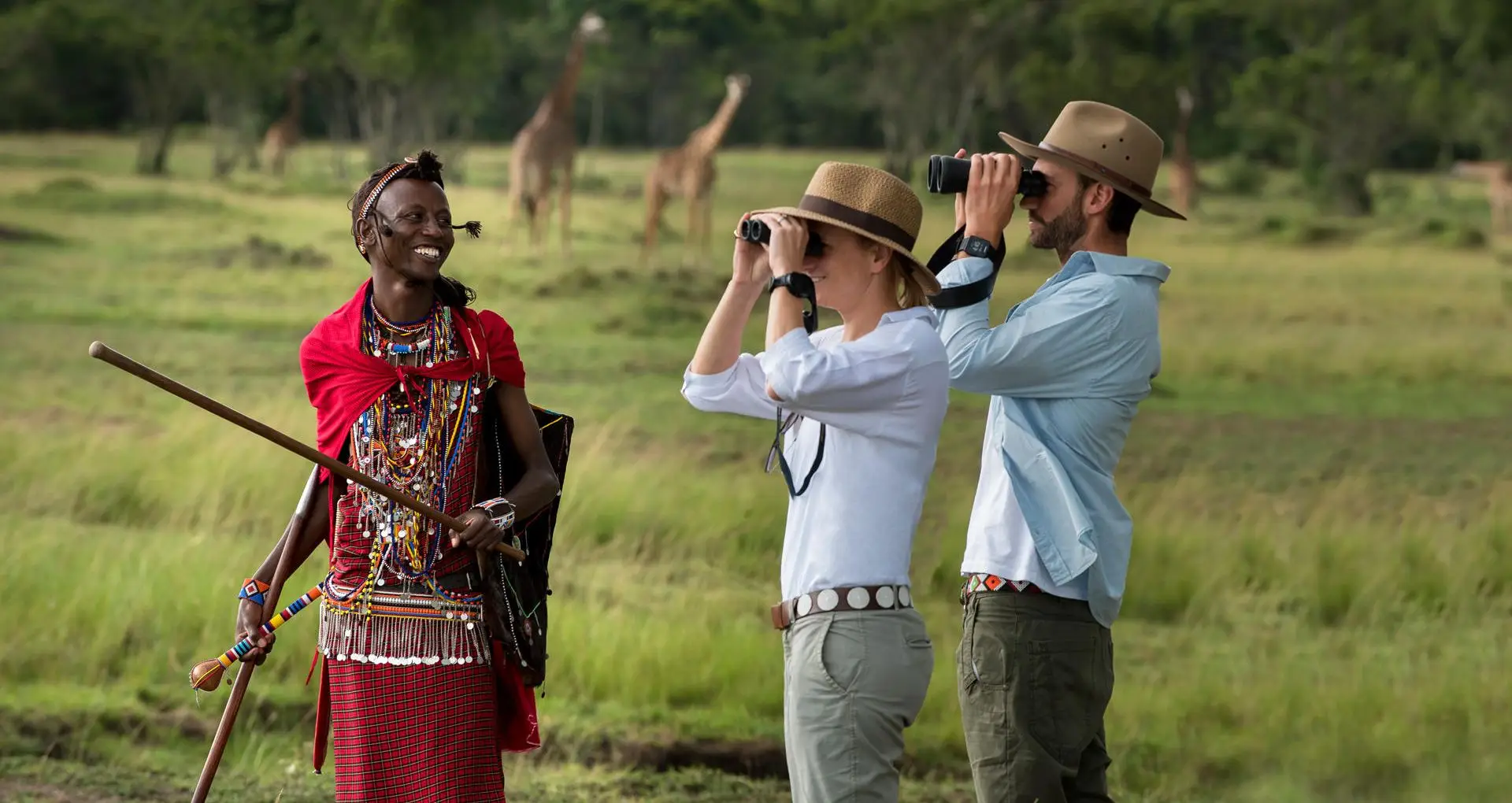 Travelers with binoculars guided by a Maasai warrior during a Safari in March, observing giraffes in the lush African savannah in Kenya