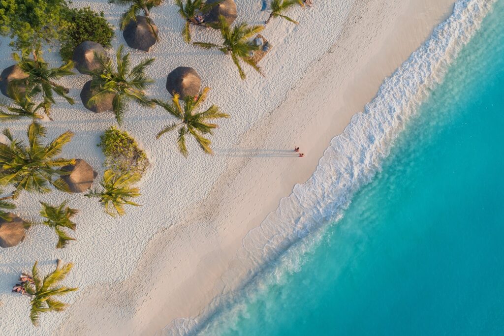 An aerial view captures a pristine white sand beach meeting turquoise ocean waves. Palm trees and thatched-roof beach umbrellas dot the sand, and two figures walk near the waterline, casting long shadows.
