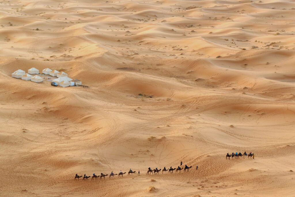 An aerial view captures a vast desert landscape with undulating sand dunes. In the distance, a cluster of white luxury tents forms a desert camp. A long line of camels, each with a rider, traverses the golden sands, evoking the adventurous spirit of Morocco Luxury Trips through the Sahara Desert.