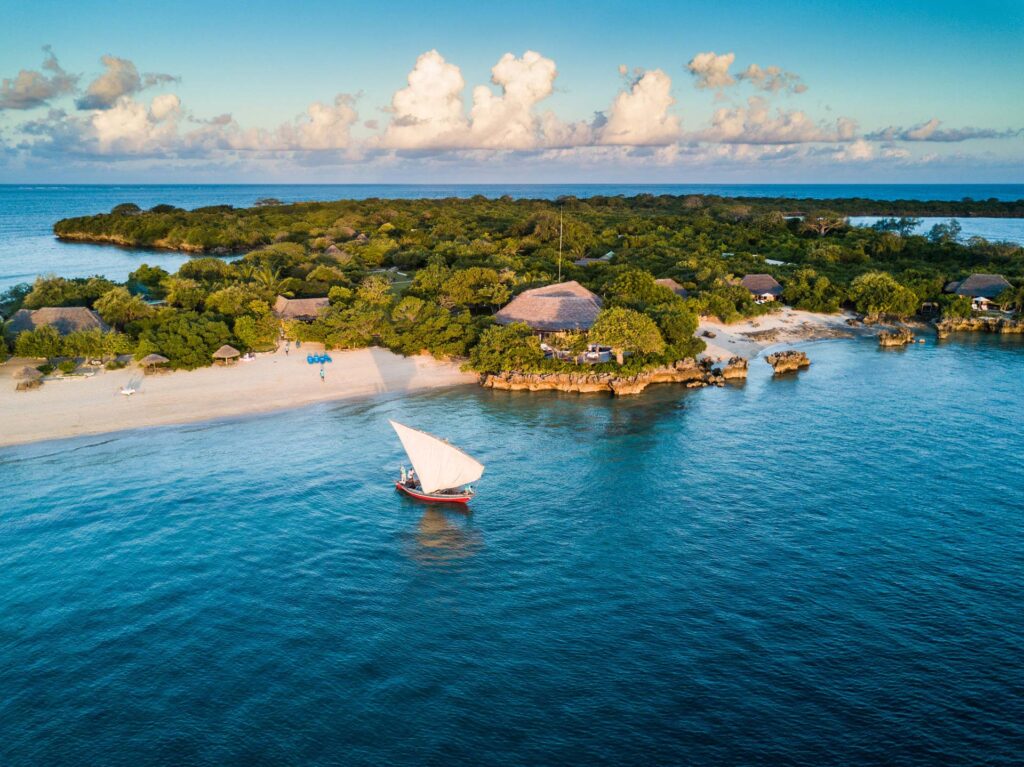 An idyllic aerial view captures a small, lush green island with white sandy beaches. Several thatched-roof bungalows are nestled among the trees. In the clear turquoise water, a traditional dhow sailboat with a white triangular sail glides gently.