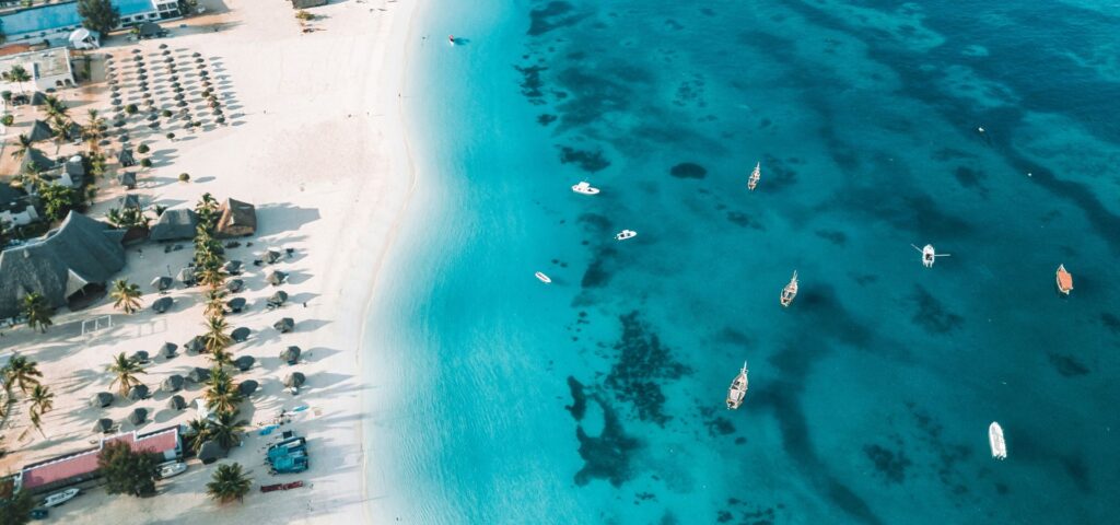 An aerial shot of a tropical beach on Zanzibar, Tanzania, showcases white sand lined with palm trees and thatched umbrellas. The brilliant turquoise ocean stretches out, dotted with numerous small boats, highlighting the serene beauty of a Zanzibar Luxury Safari.