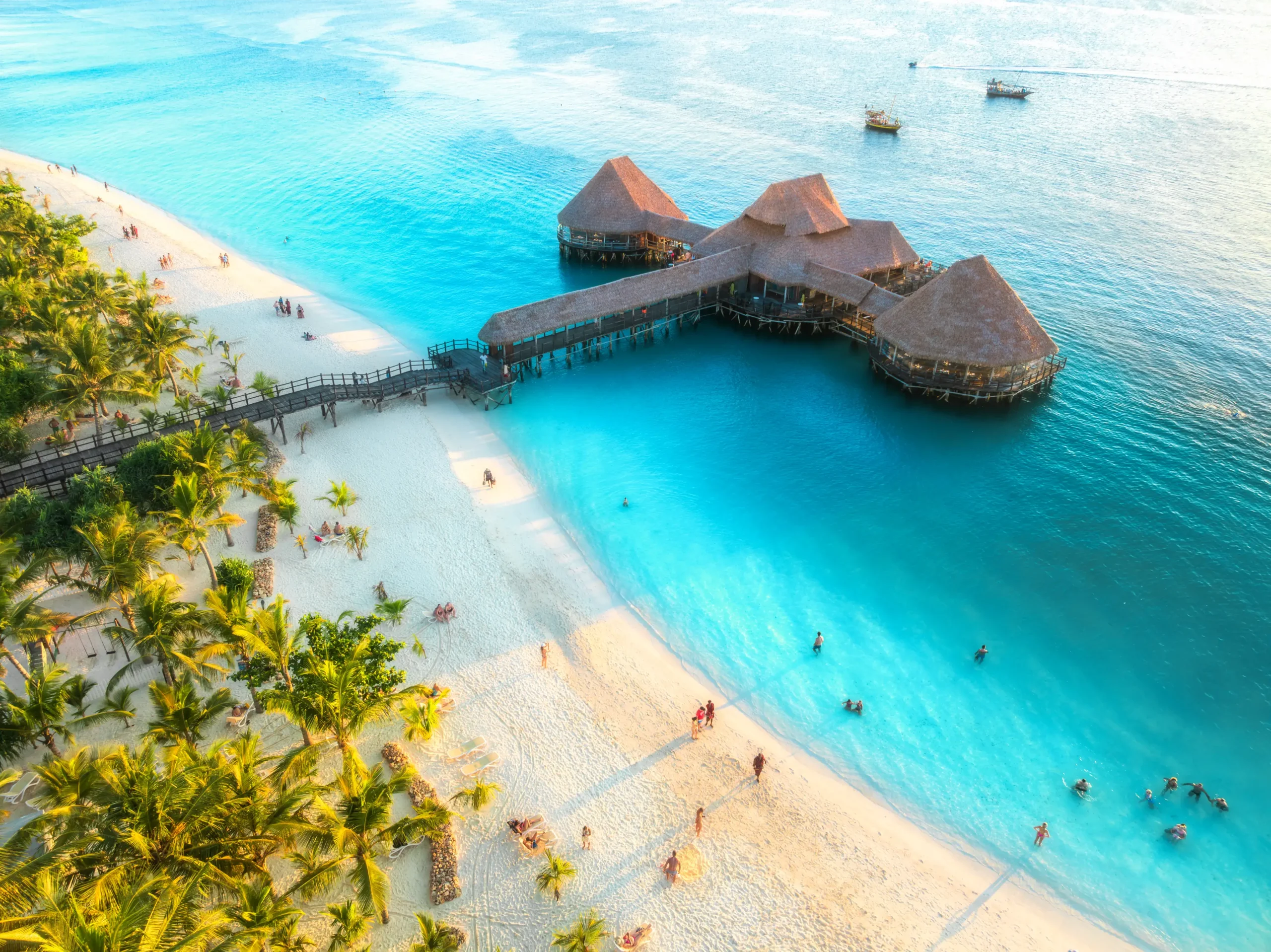 An aerial view of a wooden pier leading to a luxury thatched-roof bungalow over the vibrant turquoise waters of the Indian Ocean in Zanzibar.