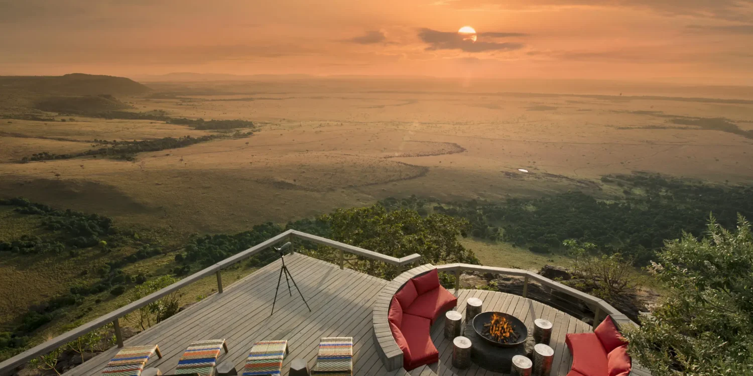 luxury outdoor lounge deck at Angama Mara at sunset overlooking a vast african savanna. the wooden deck features a circular sunken fire pit surrounded by plush red semi-circular sofas and several woven sun loungers, with a spotting scope set up to view the horizon.