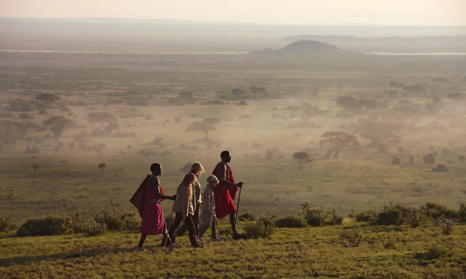 Two Maasai guides in traditional red shúkàs lead a family on a cultural walking safari through a misty landscape in East Africa.