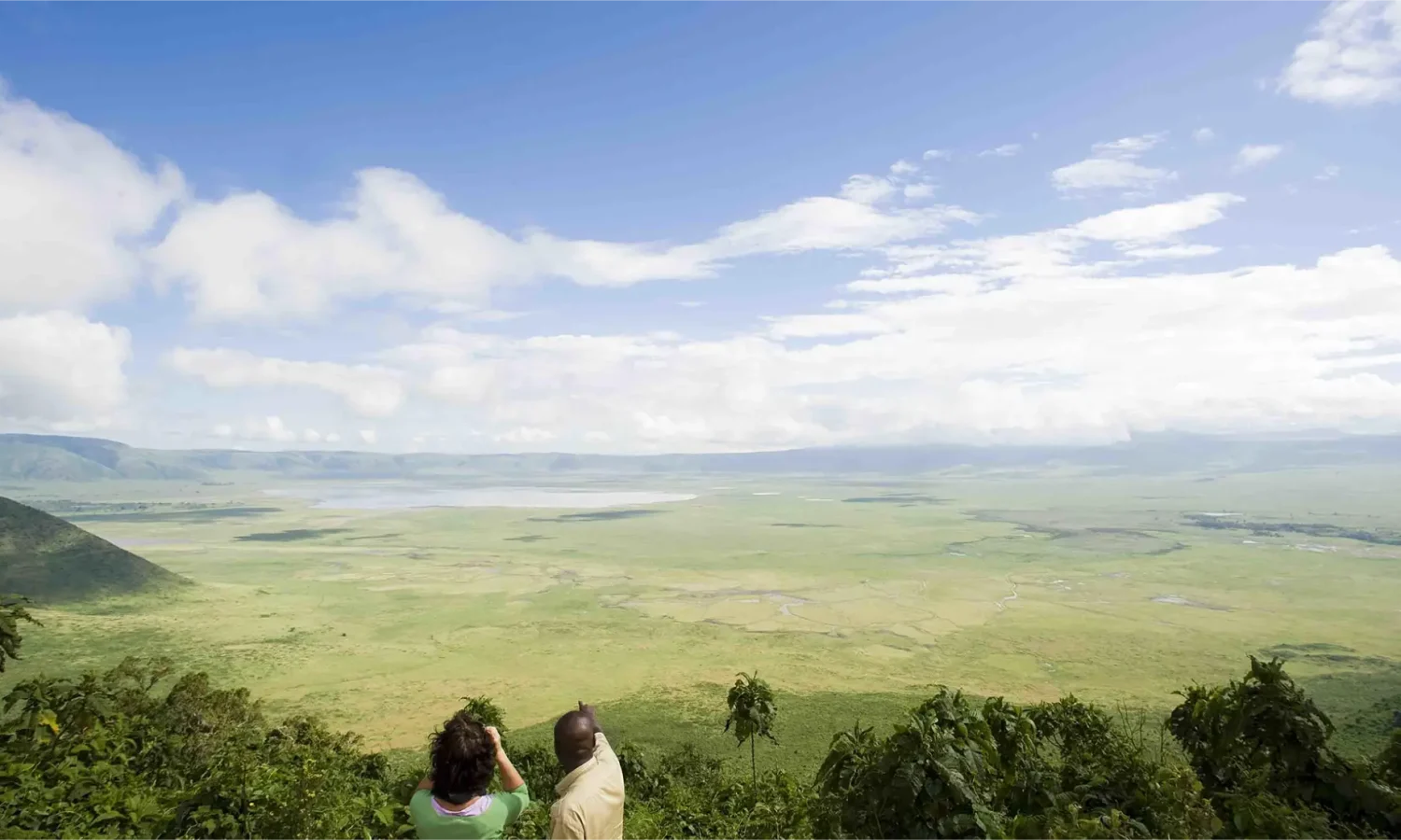 A panoramic view from a high ridge overlooking the massive, green expanse of the Ngorongoro Crater during a Tanzania safari, with two travelers looking out over the landscape.